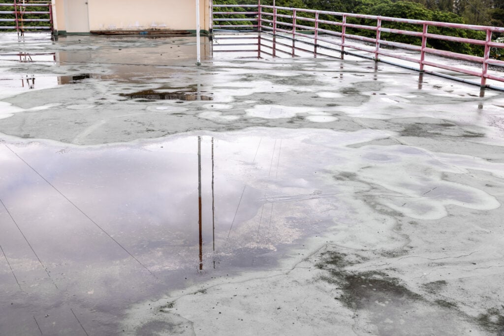 retrofit roof drains A rooftop with large puddles of water on its surface, surrounded by red metal railings. The sky and power lines are reflected in the water, and trees are visible in the background.