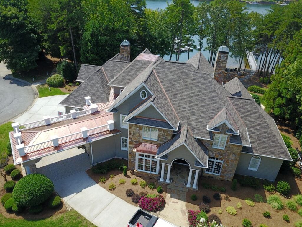 best attic ventilation A large, multi-story house with a stone and siding exterior, multiple gabled roofs, a covered driveway, and landscaped yard, situated near a lake and surrounded by trees.