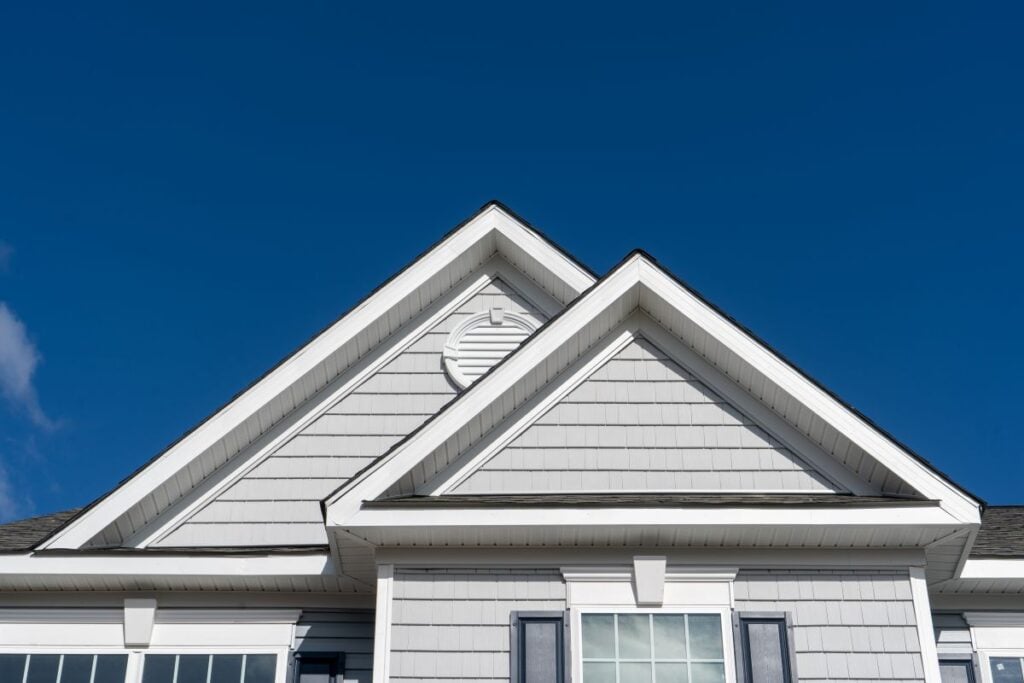 best attic ventilation The image shows the front gable of a gray house with white trim, featuring two roof peaks, double windows with dark shutters, and a bright blue sky in the background.