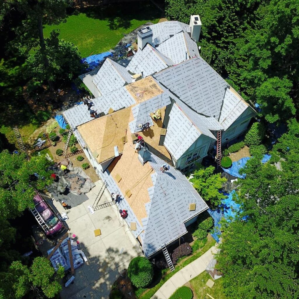 best time to replace a roof Aerial view of a house undergoing roof replacement; workers are installing new roofing materials, with sections covered in protective underlayment, ladders propped against the house, and tarps spread around the property.
