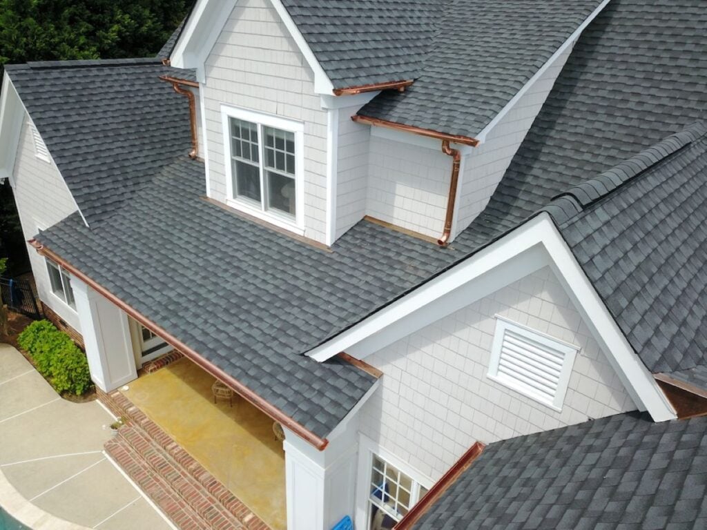 best time to replace a roof Aerial view of a house with gray asphalt shingle roofing, white siding, brick steps, and a large window, surrounded by greenery and a concrete patio area.