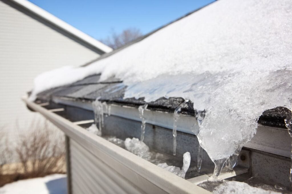 best time to replace a roof A close-up of a house roof with melting snow and ice forming icicles along the edge of the gutter on a sunny winter day. A neighboring house and bare branches are visible in the background.