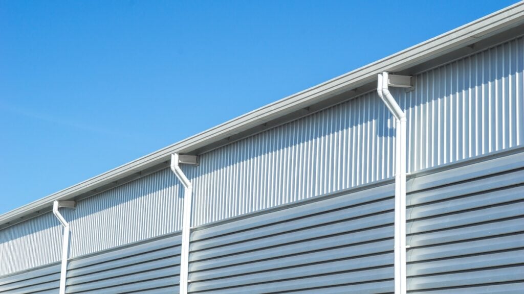 Close-up view of a modern industrial building with a corrugated metal exterior, white gutter materials, and downspouts under a clear blue sky. The structure features clean, angled lines.