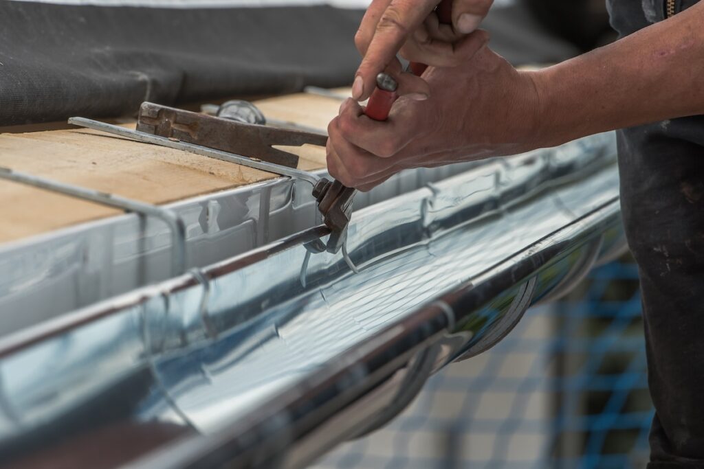 Close-up of a person using pliers to work on a shiny metal rain gutter attached to a building