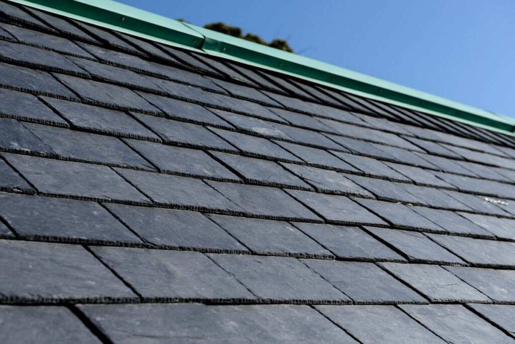 how long do tile roofs last Close-up view of a sloped roof covered with dark gray slate tiles, with a green metal ridge cap, under a clear blue sky.
