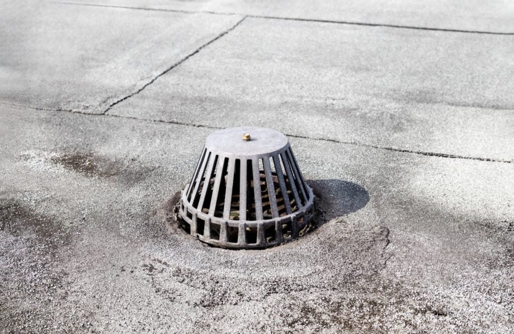 retrofit roof drains A metal roof drain cover with vertical slats sits on a flat, weathered rooftop with visible cracks and patches.