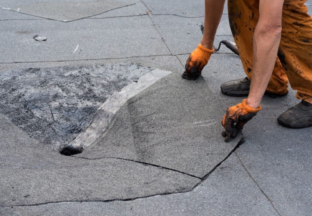 retrofit roof drains A person wearing orange gloves and work pants repairs a damaged section of a flat gray roof, lifting up part of the roofing material near a hole. Tools and tar can be seen on the surface.