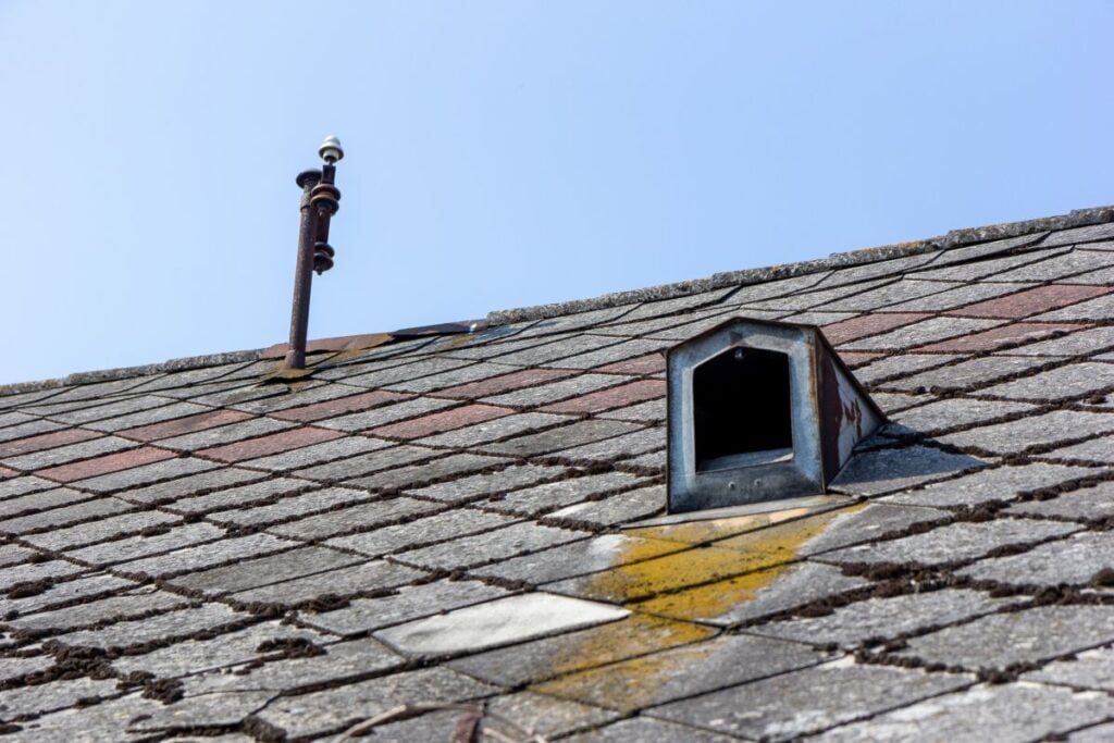 mold on roof A close-up view of a sloped, weathered shingle roof with a small, open metal vent and a metal rod with sensors or lights against a clear blue sky.