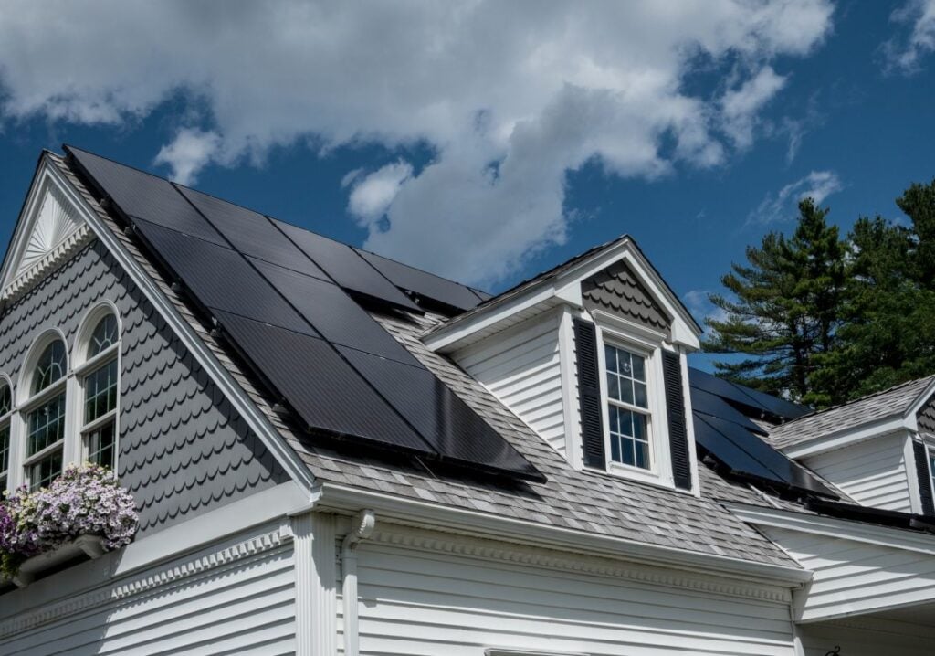 solar panel roof mounting A white house with gray shingles has multiple black solar panels installed on its roof, under a partly cloudy sky with trees in the background. Flowers hang below an arched window.