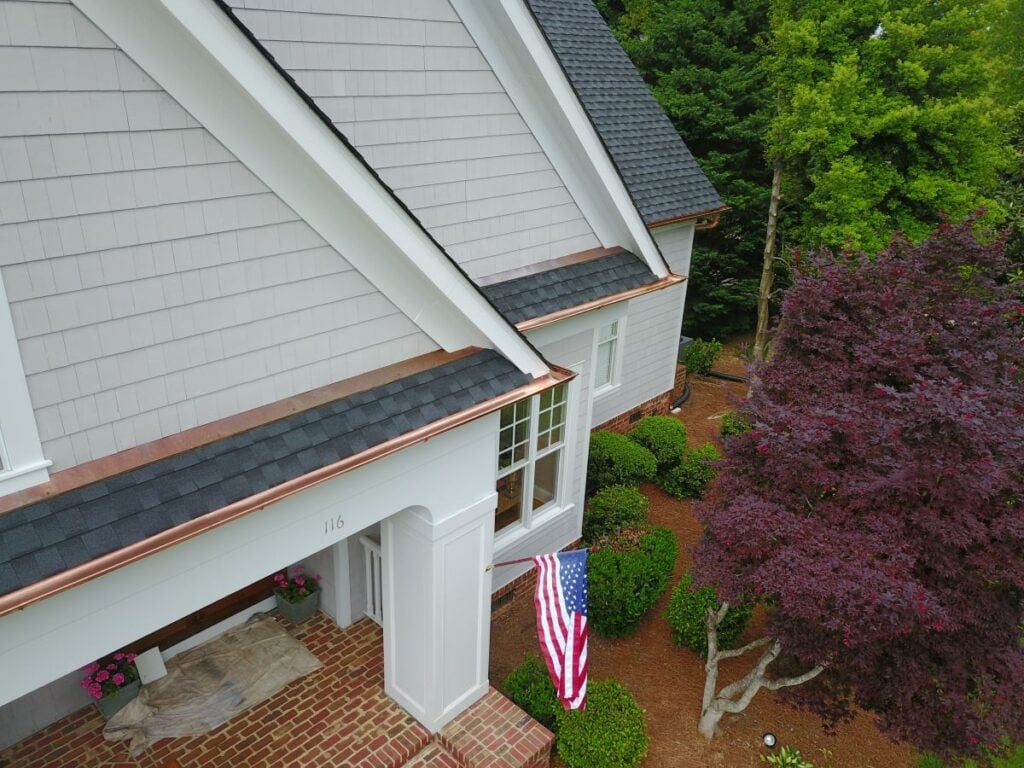 recoverable depreciation roof Aerial view of a house with gray siding, black shingle roof, and copper gutters. An American flag hangs by the front entrance, surrounded by bushes and a red-leaved tree. Brick steps lead to the doorway.