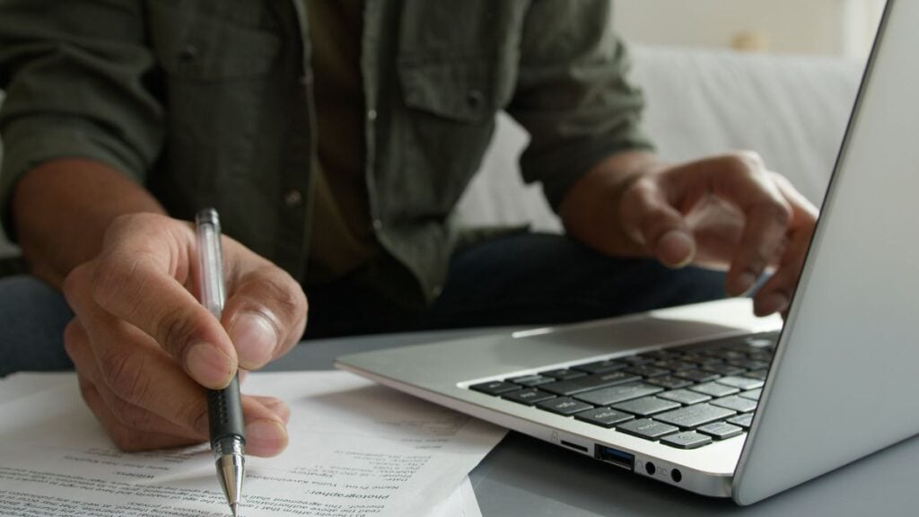 recoverable depreciation roof A person sits at a table using a laptop with one hand while writing notes on a paper with the other hand. The focus is on their hands, pen, paper, and the laptop keyboard.