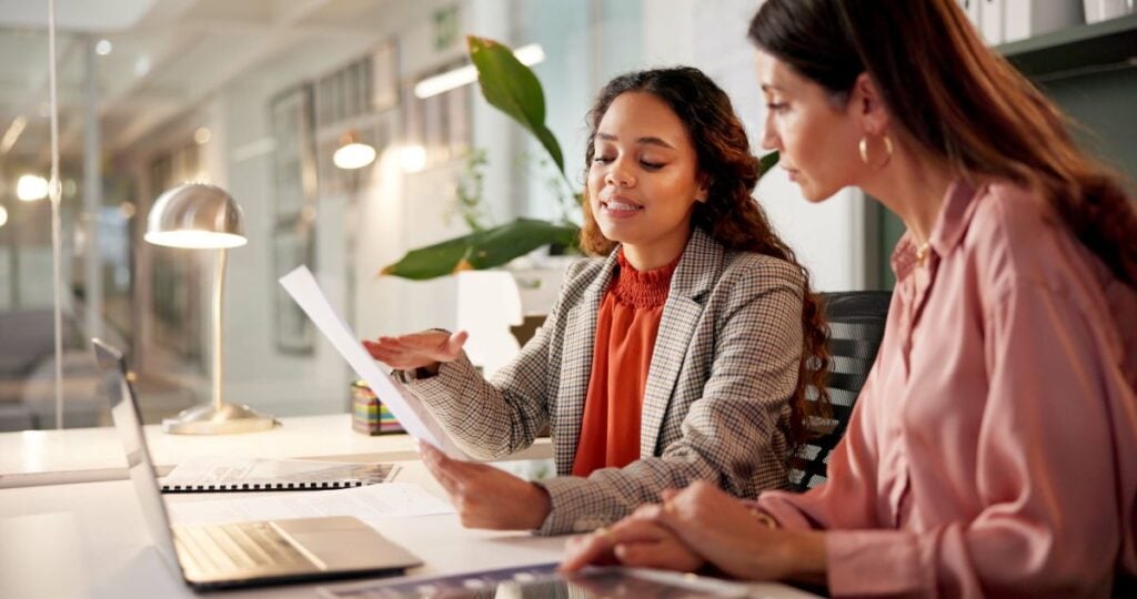 recoverable depreciation roof Two women sitting at a desk in an office, reviewing documents together. One woman is holding a paper and explaining something, while the other listens attentively. A laptop and lamp are on the desk.