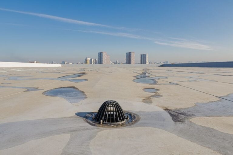 retrofit roof drains A flat white rooftop with scattered puddles, featuring a central drain in the foreground; tall buildings and a clear blue sky are visible in the distant background.