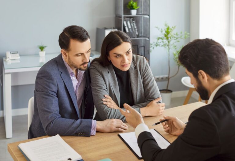 recoverable depreciation roof A couple sits closely together at a desk, listening seriously to a man in a suit who is explaining documents to them in a bright office setting.