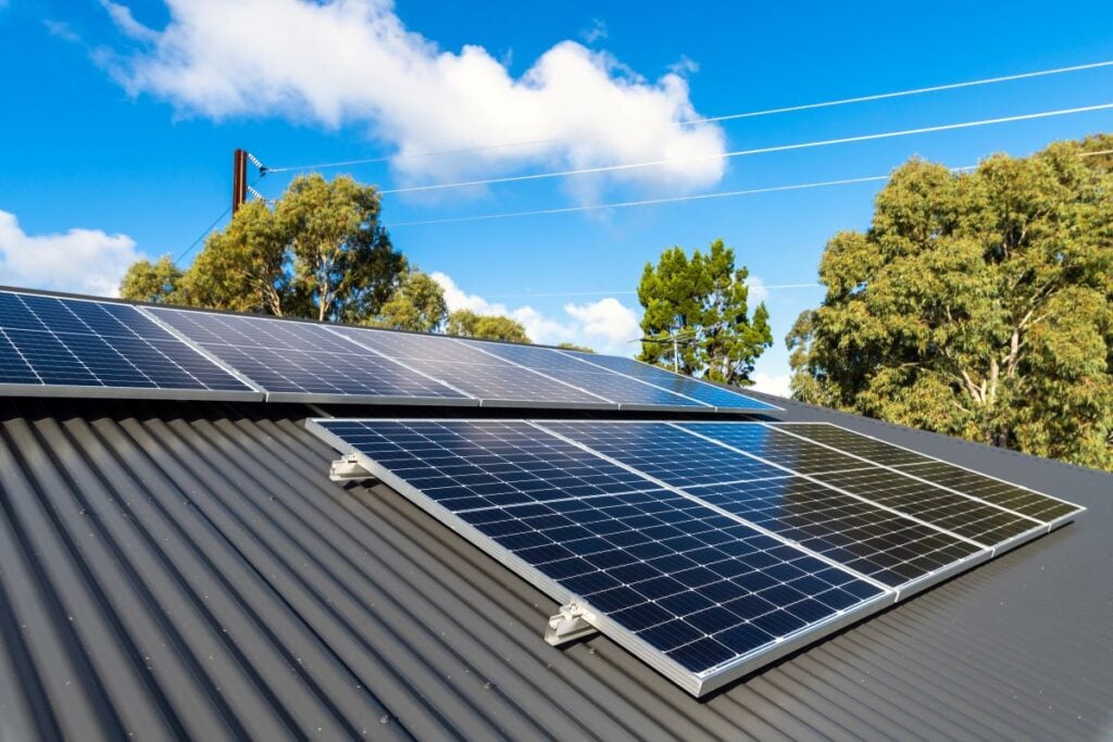 solar panel roof mounting Solar panels installed on a corrugated metal roof, with trees and power lines in the background under a blue sky with scattered clouds.
