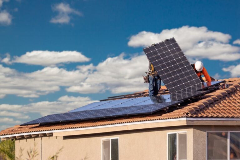 solar panel roof mounting Two workers in safety gear install large solar panels on a tiled residential roof under a blue sky with scattered clouds.