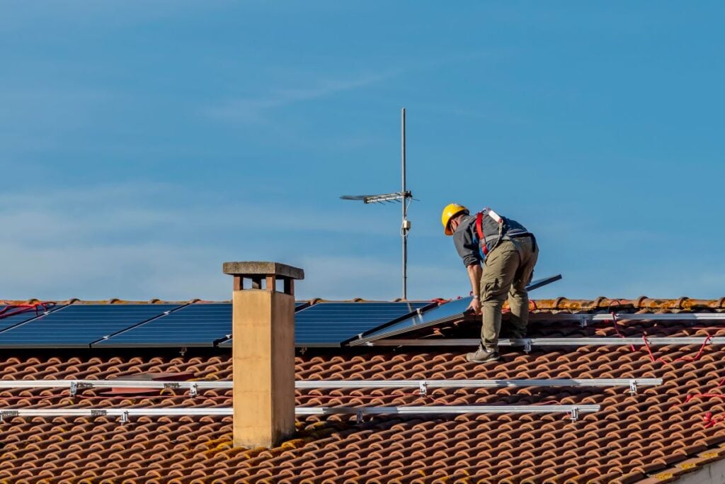 solar panel roof mounting A worker wearing a yellow helmet and safety harness installs solar panels on a tiled roof under a clear blue sky. An antenna and chimney are also visible on the roof.