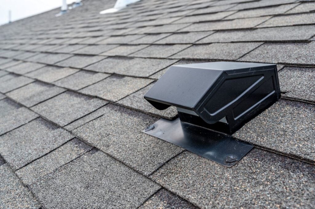 best attic ventilation Close-up of a black roof vent installed on an asphalt shingle roof, with overlapping gray shingles visible in the background.