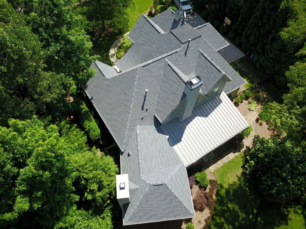 Aerial view of a large house with a complex, dark gray shingle roof surrounded by green trees and landscaped gardens on a sunny day. A white vehicle is parked in the driveway.