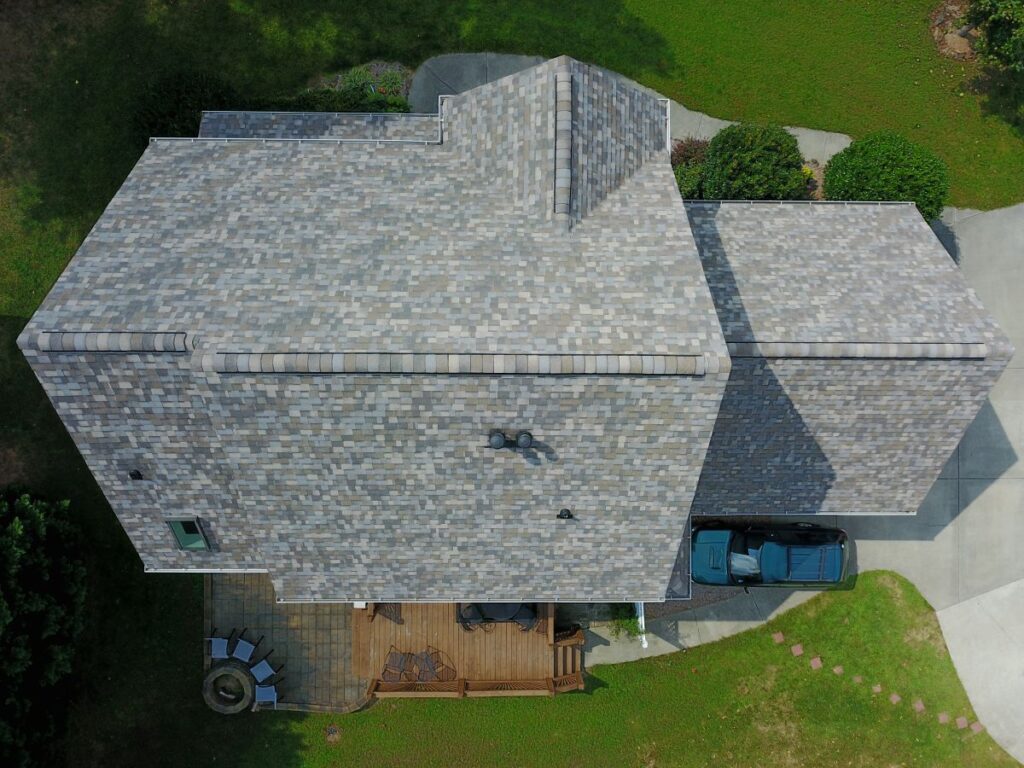 Aerial view of a house with gray shingle roof, wooden backyard deck with patio furniture, two cars parked in the driveway, and green lawn surrounding the home.
