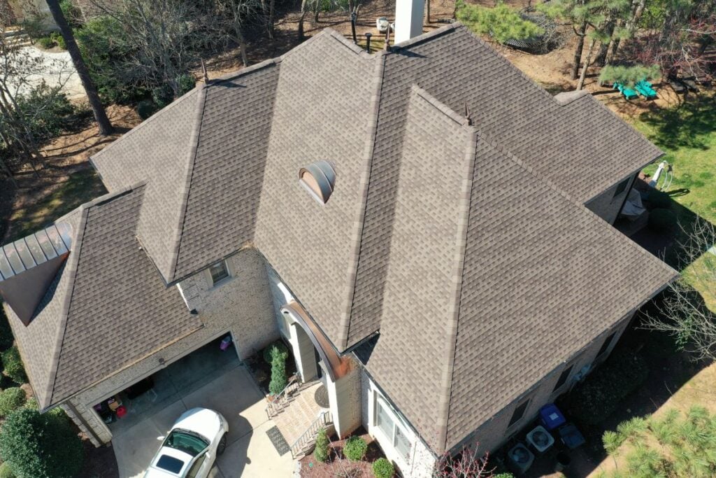 Aerial view of a large house with a complex, multi-gabled brown shingle roof, brick exterior, driveway, white car, and surrounding trees and shrubs.