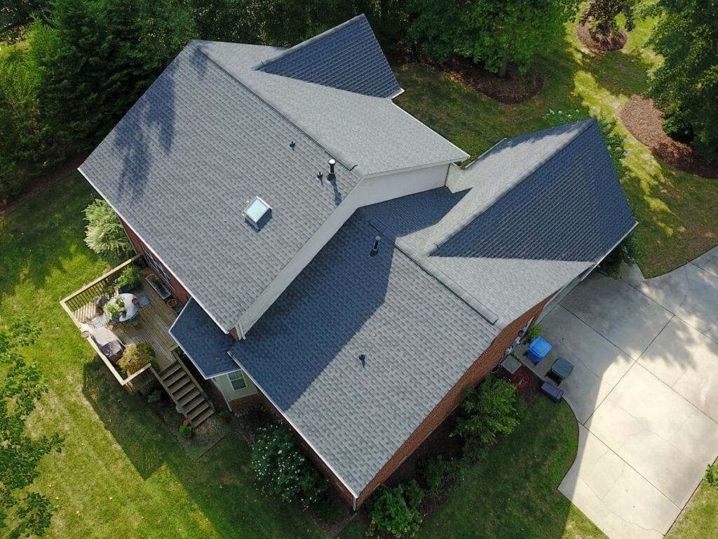 Aerial view of a house with a gray shingle roof, attached wooden deck with outdoor furniture, curved driveway, green lawn, and surrounding trees and shrubs.