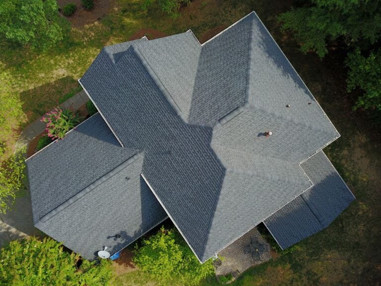 Aerial view of a gray-shingled house roof surrounded by green trees and patches of grass, with walkways and a small patio visible around the building.