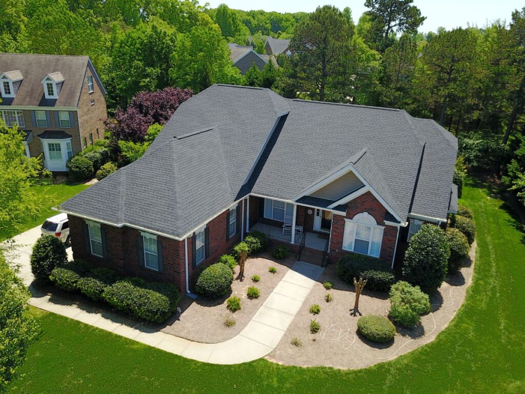 Aerial view of a large, single-story brick house with a dark gray roof, surrounded by green lawns, bushes, and trees in a suburban neighborhood on a sunny day.