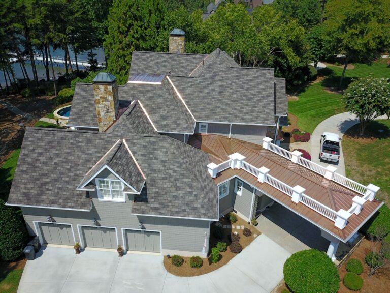 Aerial view of a large suburban house with gray shingles, three-car garage, two stone chimneys, and a raised walkway leading to the upper level. The property is surrounded by trees, lawn, and a white car in the driveway.
