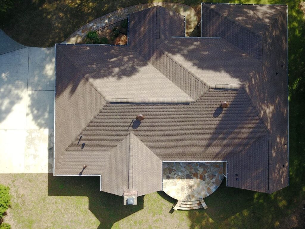Aerial view of a house with a brown shingle roof, stone patio, and surrounding green lawn. Shadows of trees are visible on the roof, and a driveway is on the left side of the image.
