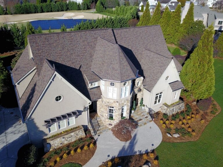 Aerial view of a large, two-story house with a stone entrance, beige walls, and a dark shingled roof, surrounded by landscaped gardens, trees, and a well-manicured lawn. A golf course with a pond is visible in the background.