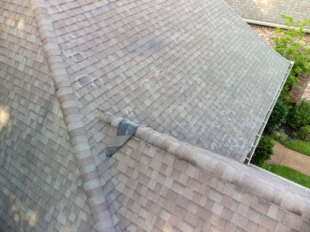 A house roof with light brown shingles shows numerous chalk circles marking spots, likely indicating areas of hail or storm damage. Trees and a portion of a brick wall are visible in the background.