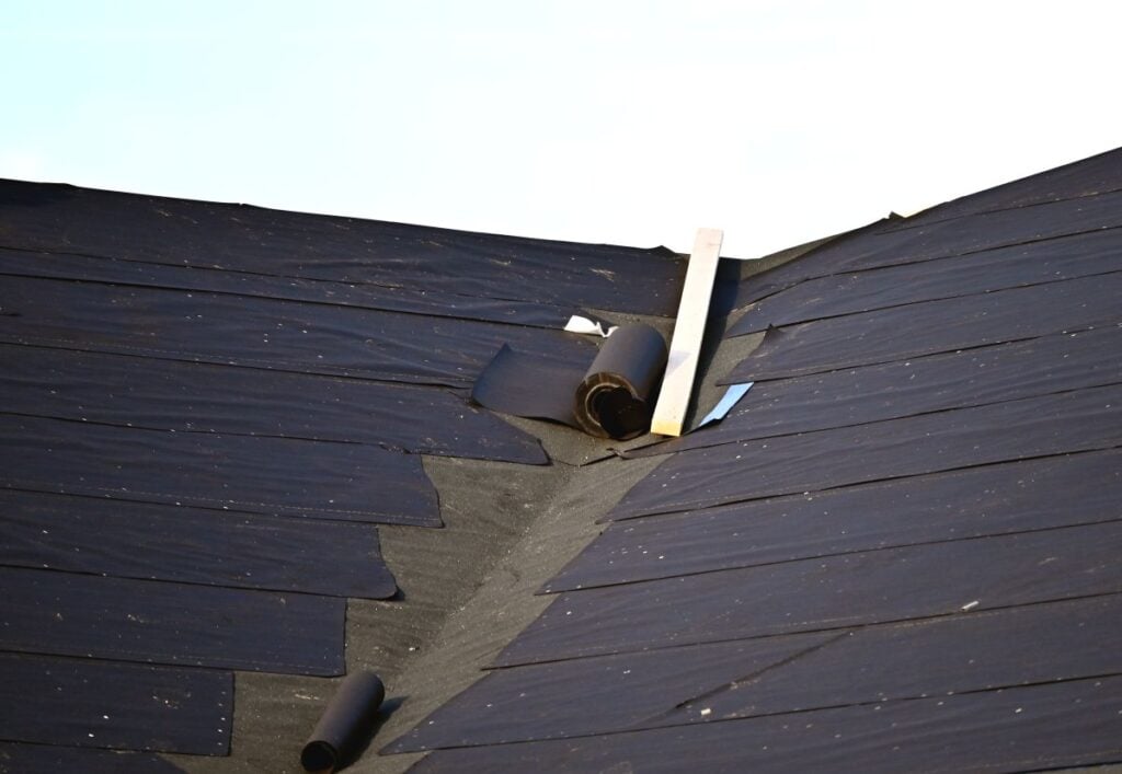 A roof under construction with black roofing felt partially installed; rolls of felt and a wooden plank rest near the valley where two roof sections meet. The sky is clear in the background.