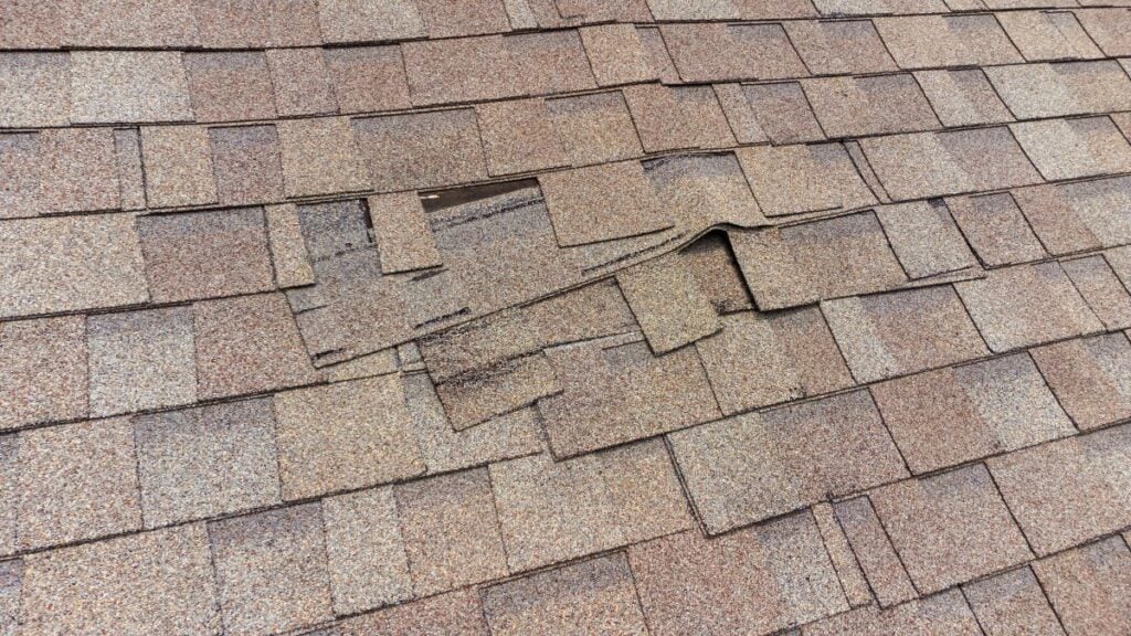 Asphalt roof with several shingles lifted and dislodged, exposing the underlying layer and creating uneven, raised sections in the middle of the roof.