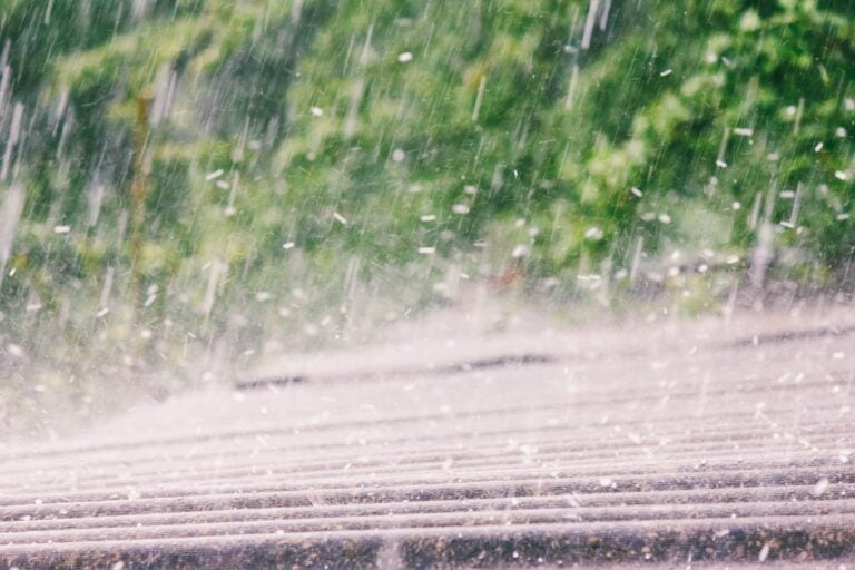 Heavy rain falls on a slanted rooftop with blurred green foliage in the background, creating a sense of movement and wet weather.