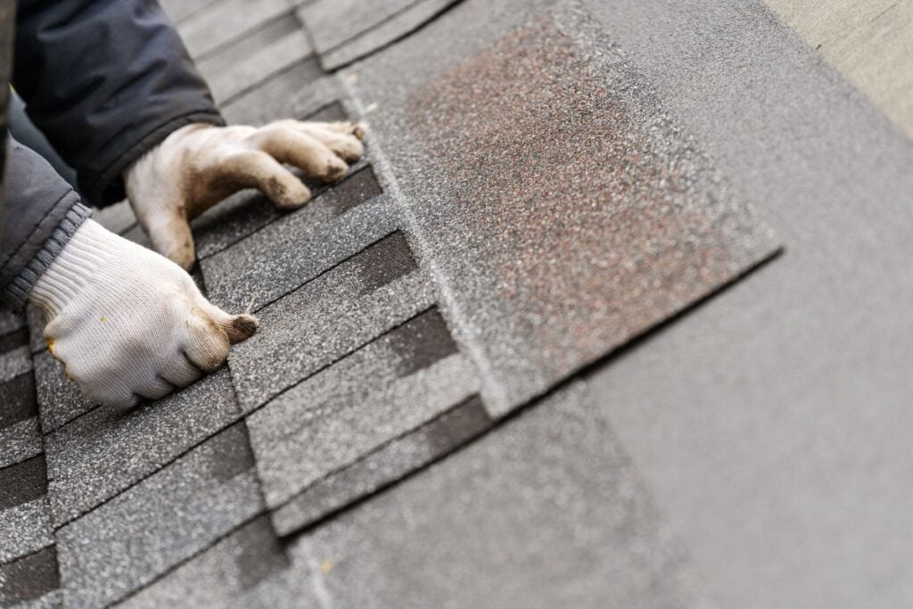 A person wearing white work gloves is installing asphalt shingles on a roof, carefully aligning each shingle for proper placement. The close-up shows the hands and the textured roof surface.