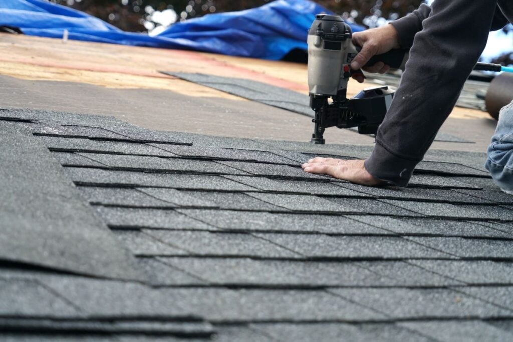 A person installs asphalt shingles on a roof using a nail gun. The individual is kneeling, with one hand steadying the shingles and the other operating the tool. Roofing materials and a blue tarp are visible in the background.