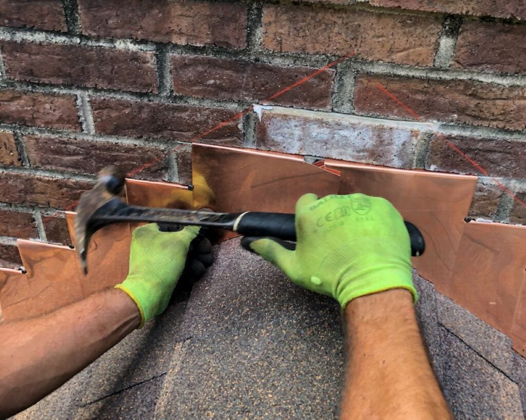 roof to wall flashing detail A person wearing green gloves uses a hammer to install copper roof to wall flashing detail on a shingled roof beside a brick wall. Red chalk lines are visible on the wall for alignment.