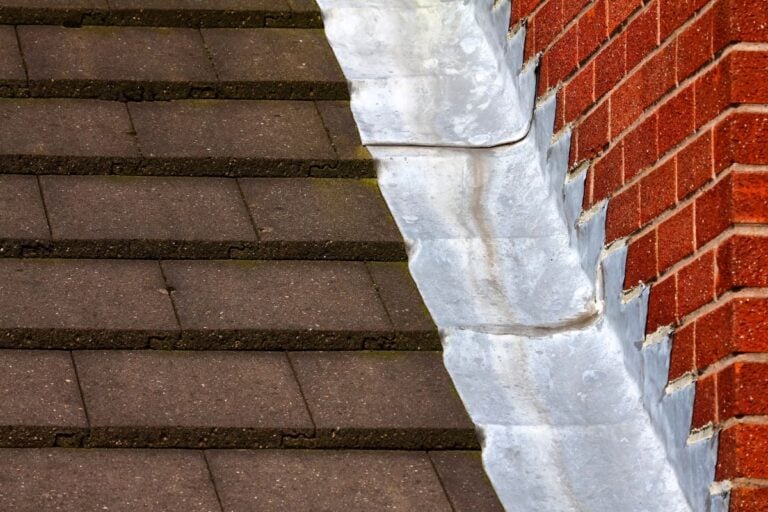 roof to wall flashing detail Close-up of a roof to wall flashing detail, where brown shingles meet a red brick wall, with a strip of metal flashing installed at the intersection for waterproofing.