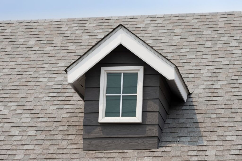 what is a gaf master elite contractor A small dormer window with white trim extends from a gray shingled roof. The window has four panes and is framed by dark gray siding. The sky is slightly visible in the background.