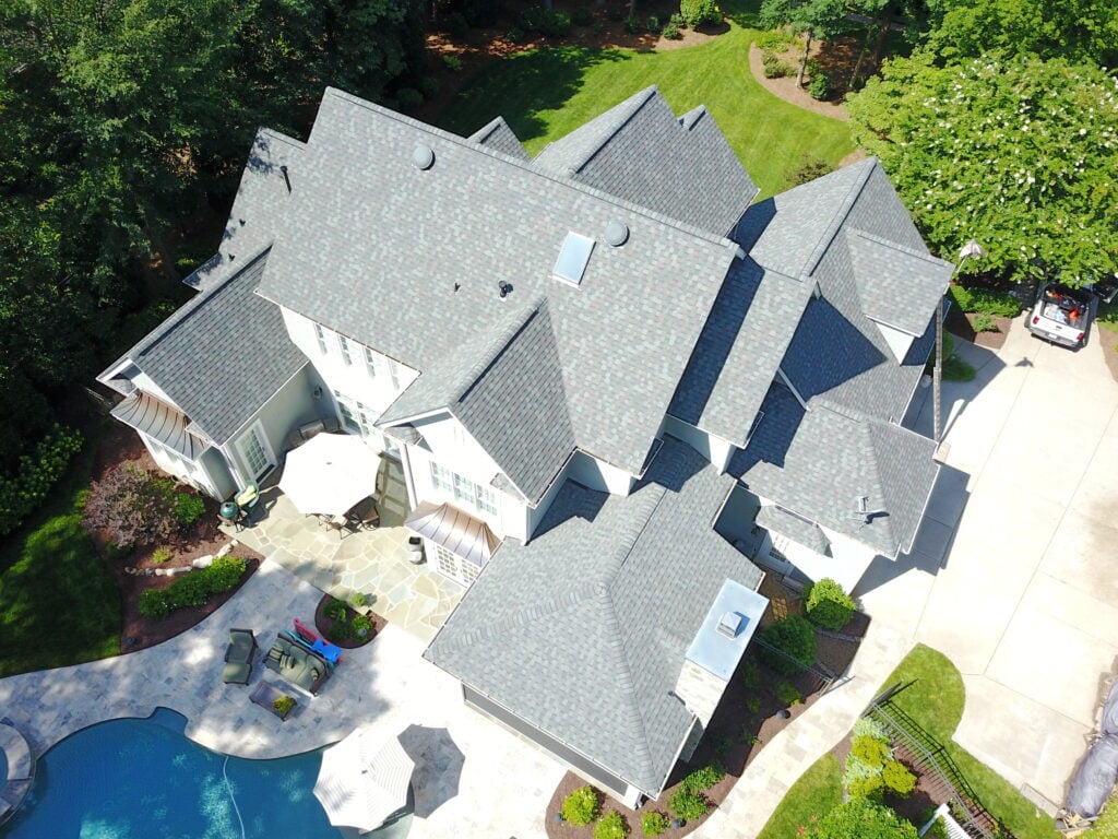 roof replacement cost charlotte nc Aerial view of a large house with gray roofing, a backyard pool, patio furniture, landscaped garden, and a driveway with a white car parked near the garage.