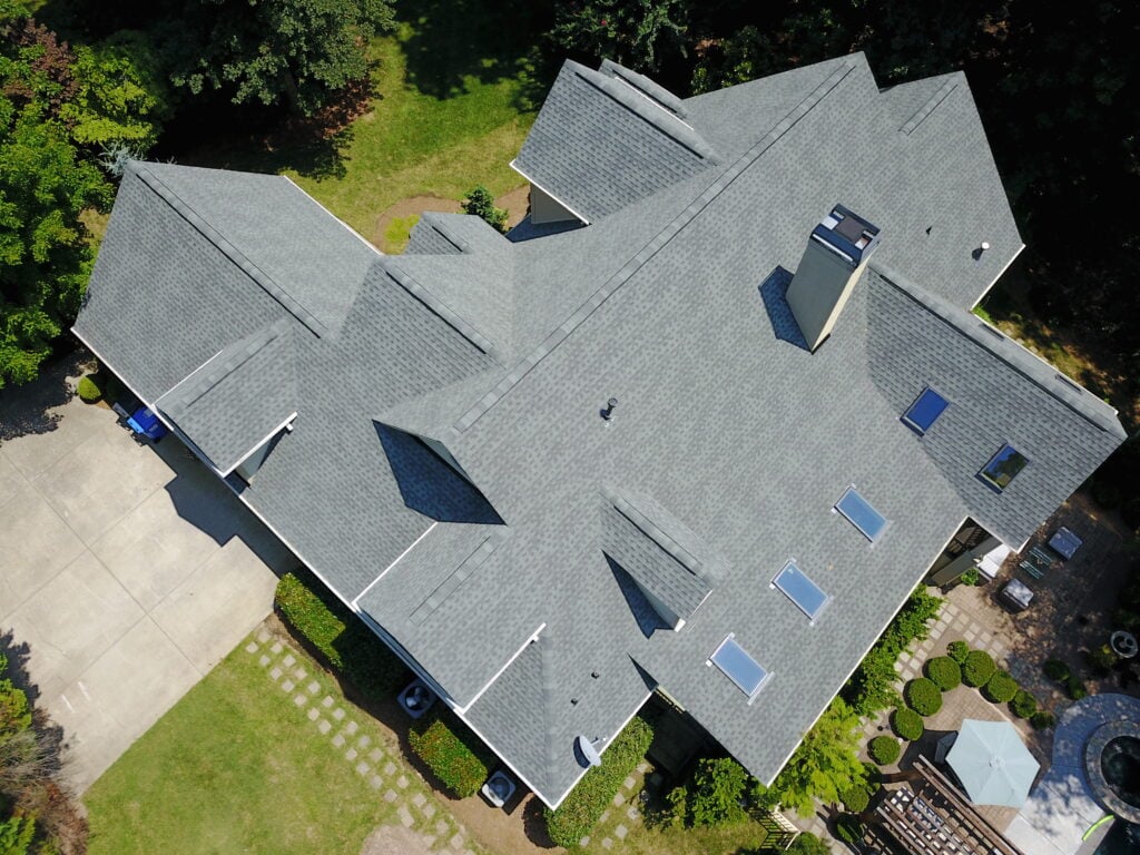 roof replacement cost raleigh nc Aerial view of a large gray-roofed house with skylights, surrounded by greenery, a driveway, and a patio area with outdoor furniture.