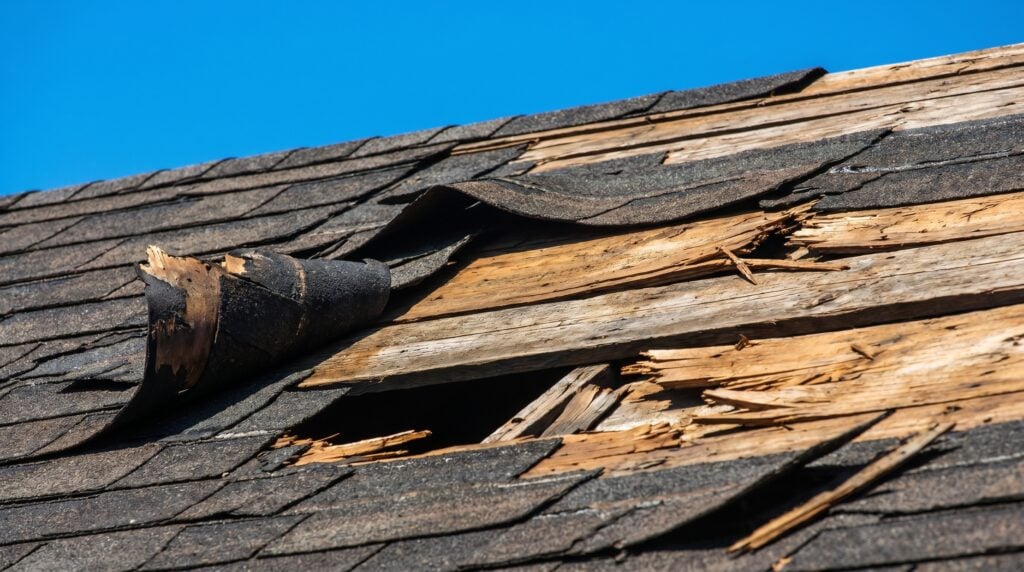 best roofing materials north carolina Damaged roof with broken and missing shingles, revealing splintered wooden boards underneath against a clear blue sky.
