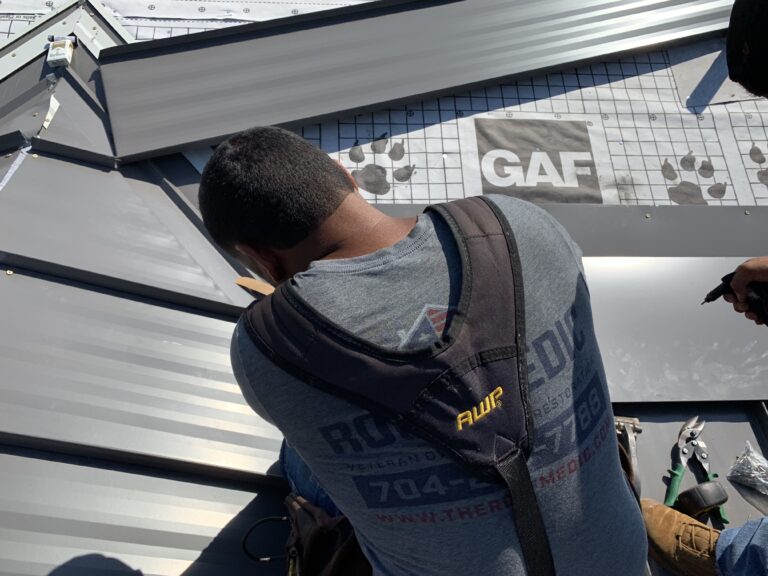 roof replacement cost charlotte nc A worker in a harness installs metal roofing panels on a roof. Roofing underlayment with a GAF logo is visible, along with tools and sunlit panels.