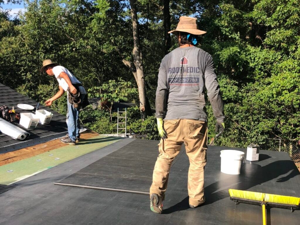 roof replacement cost charlotte nc workers standing on top of shingles roof Two workers in hats and long sleeves install roofing material on a flat roof on a sunny day. One stands near a broom, while the other handles white buckets. Trees are visible in the background.
