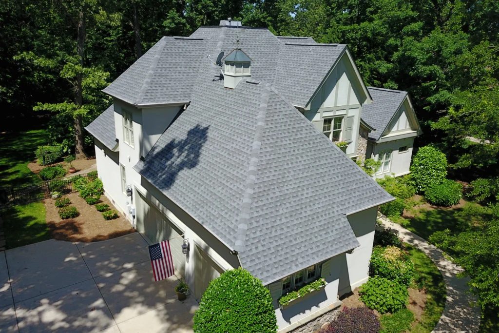 best roofing materials north carolina Aerial view of a large white house with a gray shingle roof, surrounded by green trees, bushes, and a driveway with an American flag displayed near the entrance.