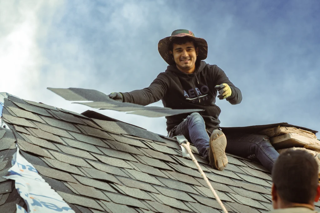 what is a gaf master elite contractor A smiling person wearing a hat and hoodie installs shingles on a rooftop under a blue sky. They sit near the roof’s peak, holding a shingle, with a rope secured nearby.