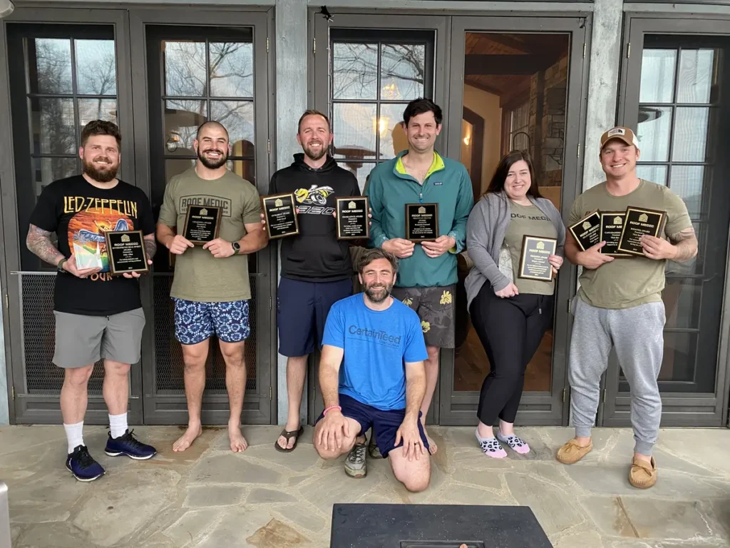 Seven people stand on a stone patio holding award plaques, smiling for the camera in front of glass doors. One person kneels in front, while the rest stand close together in casual clothing, celebrating recognition for their outstanding work in Charlotte roof replacement.