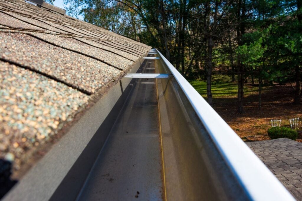 Close-up of a clean, empty rain gutter attached to the edge of a shingled roof. Trees and yard are visible in the blurred background on a sunny day.