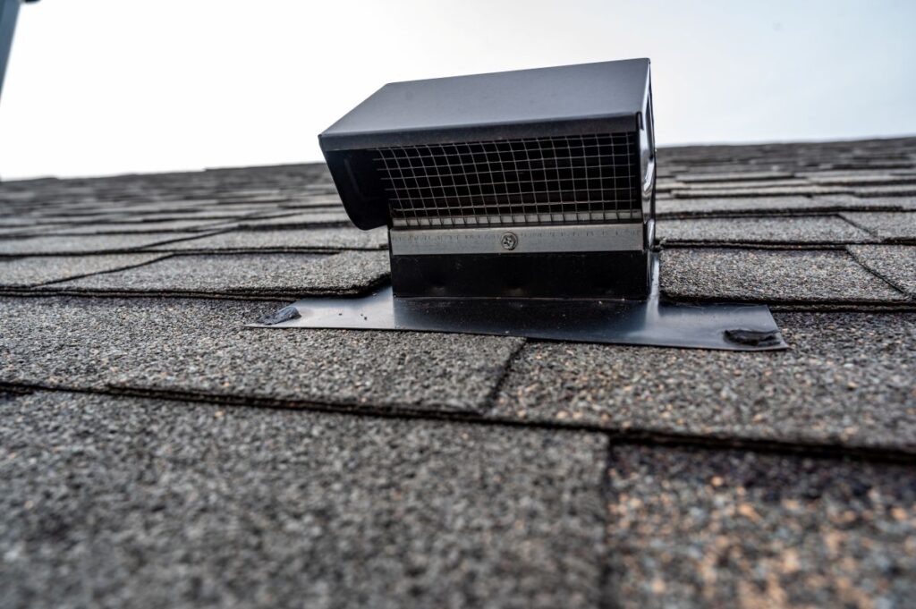 A close-up view of a black roof vent with a wire mesh cover, installed on an asphalt shingle roof against a light sky background.